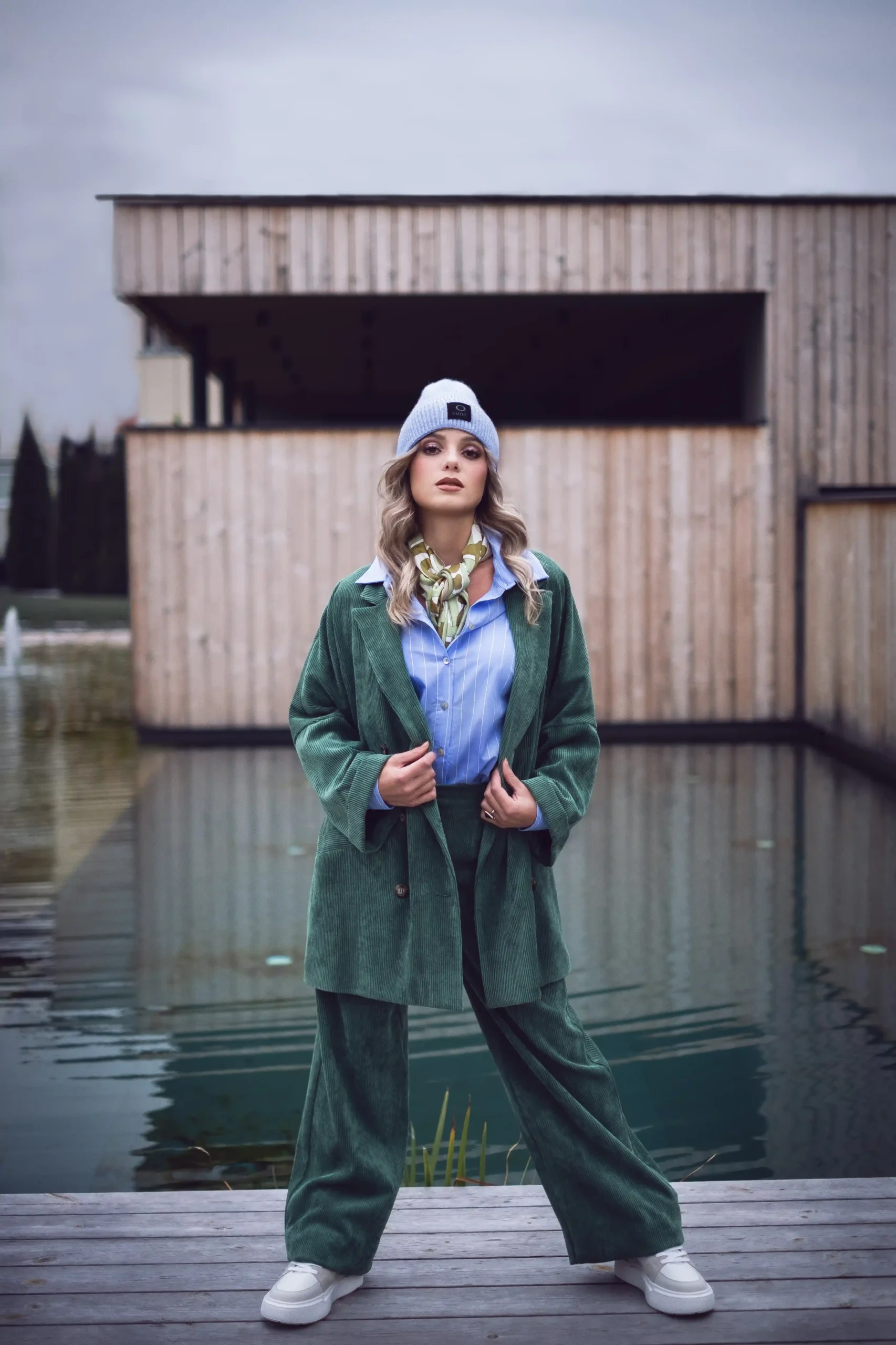 Una donna si trova in piedi con sicurezza su un ponte di legno sopra l'acqua, indossando un abito di velluto a coste verde e un berretto grigio, mentre moderni edifici in legno incorniciano la scena, ripresi con produzione foto e video professionale per un effetto visivo di grande impatto.