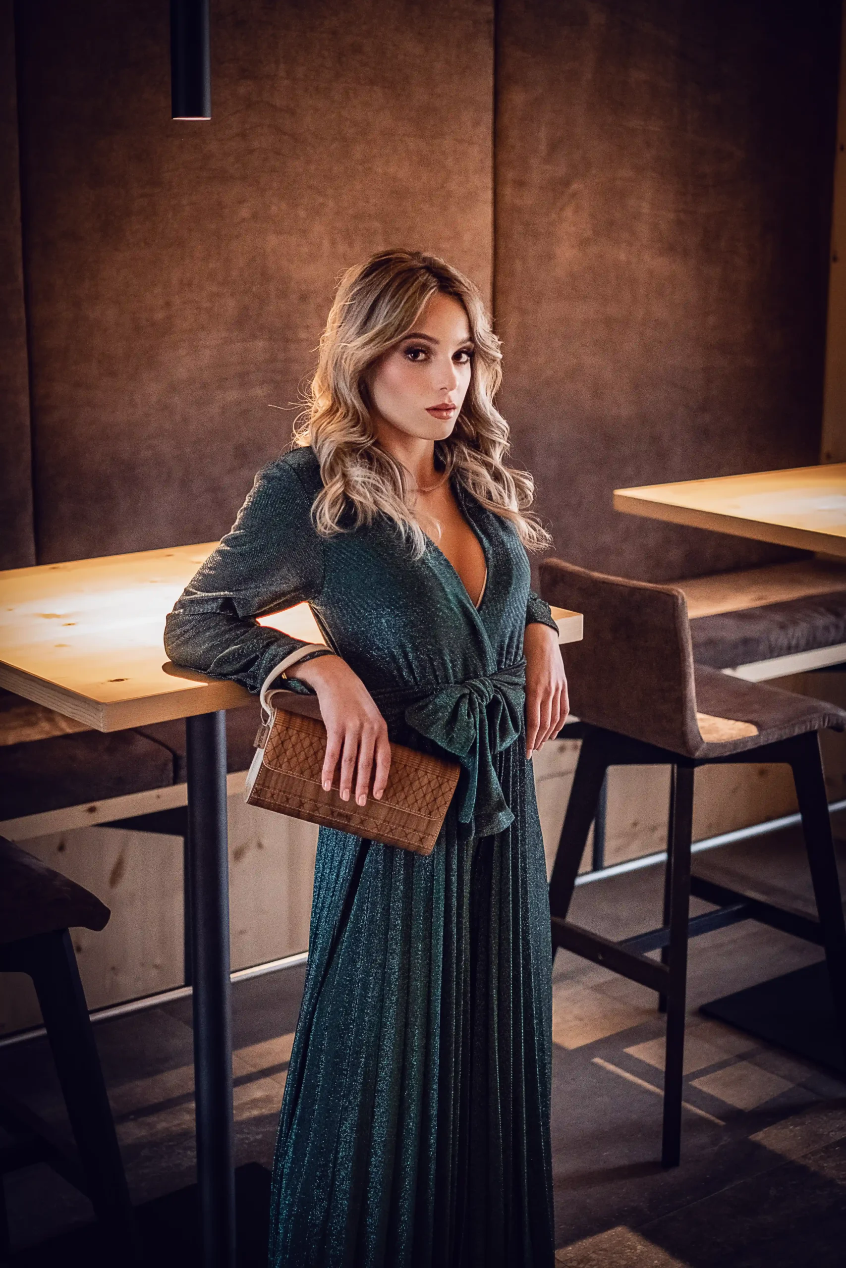 Una donna con i capelli biondi ondulati che indossa un abito verde scuro scintillante sta in piedi con sicurezza accanto a un tavolo alto, tenendo in mano una pochette marrone, in un ristorante moderno poco illuminato con accenti in legno, perfetto per la produzione di foto e video professionali.