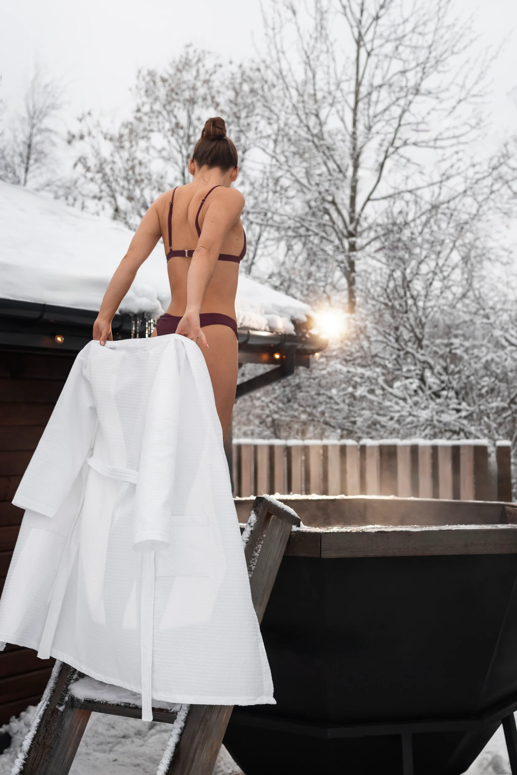 Una donna in bikini tiene in mano un accappatoio bianco mentre sale in una vasca idromassaggio all'aperto circondata da neve e alberi: una scena straordinaria che cattura la produzione foto e video professionale in un pittoresco ambiente invernale.