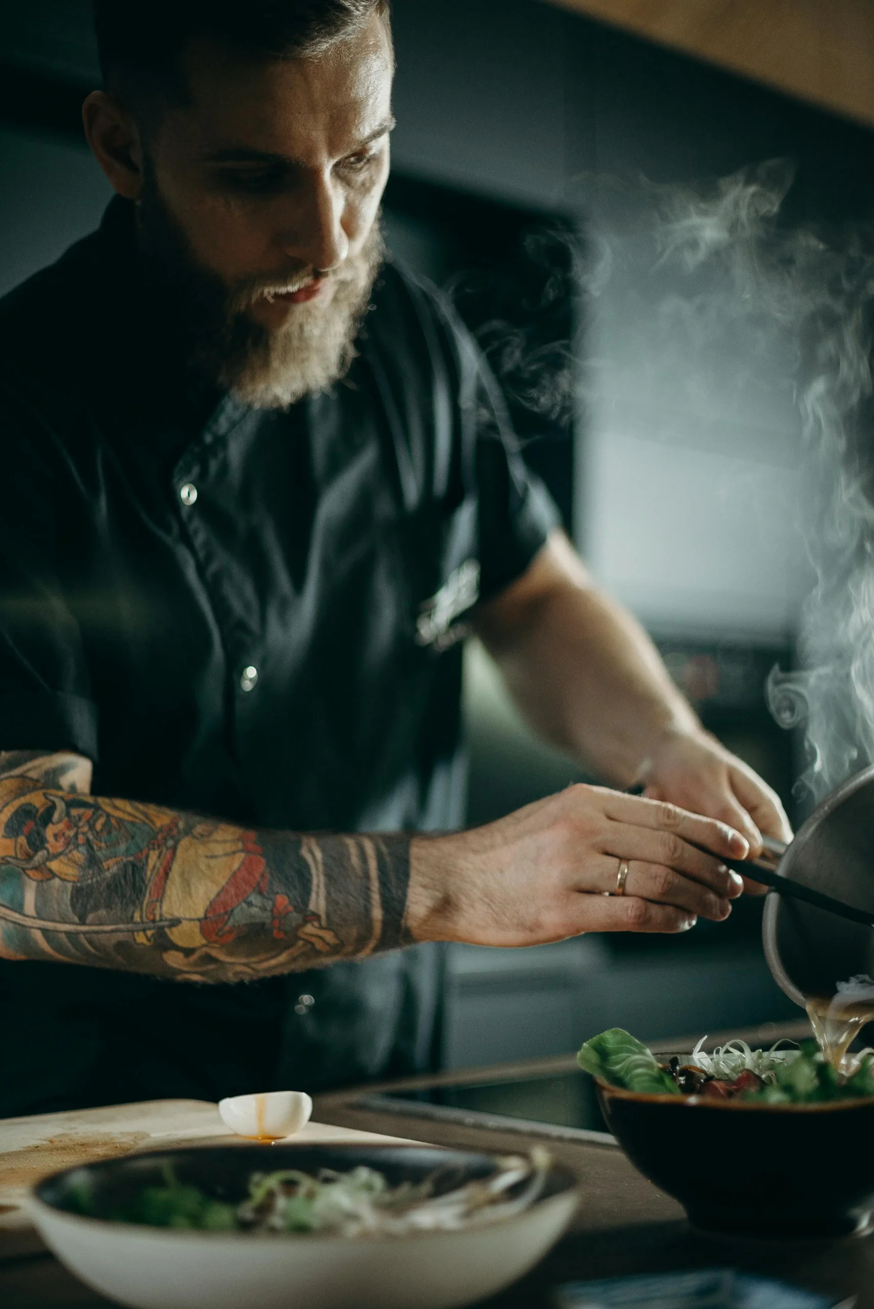 Uno chef con barba e braccio tatuato, in uniforme nera, versa cibo fumante da una pentola su un piatto in una cucina durante una produzione foto e video professionale.