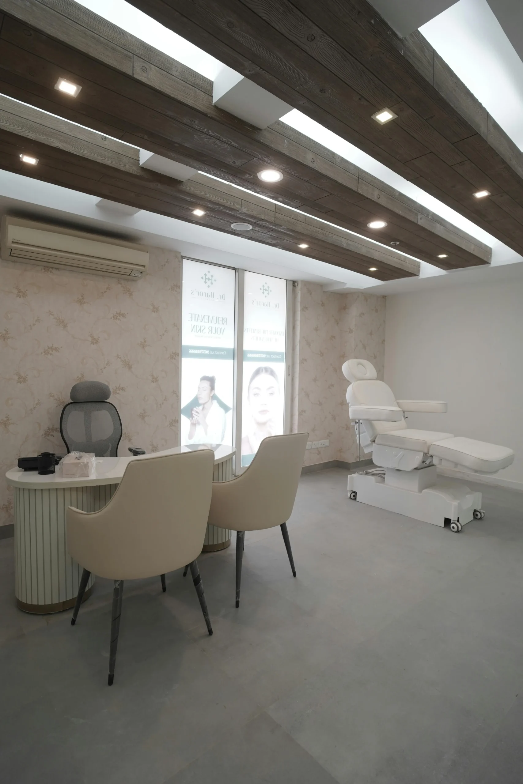 Un ambulatorio moderno con una scrivania bianca, due sedie beige, una poltrona da ufficio e un lettino da visita reclinabile. Grandi finestre con poster di bellezza lasciano entrare la luce naturale; la stanza presenta travi in legno sul soffitto e carta da parati a fiori chiari.