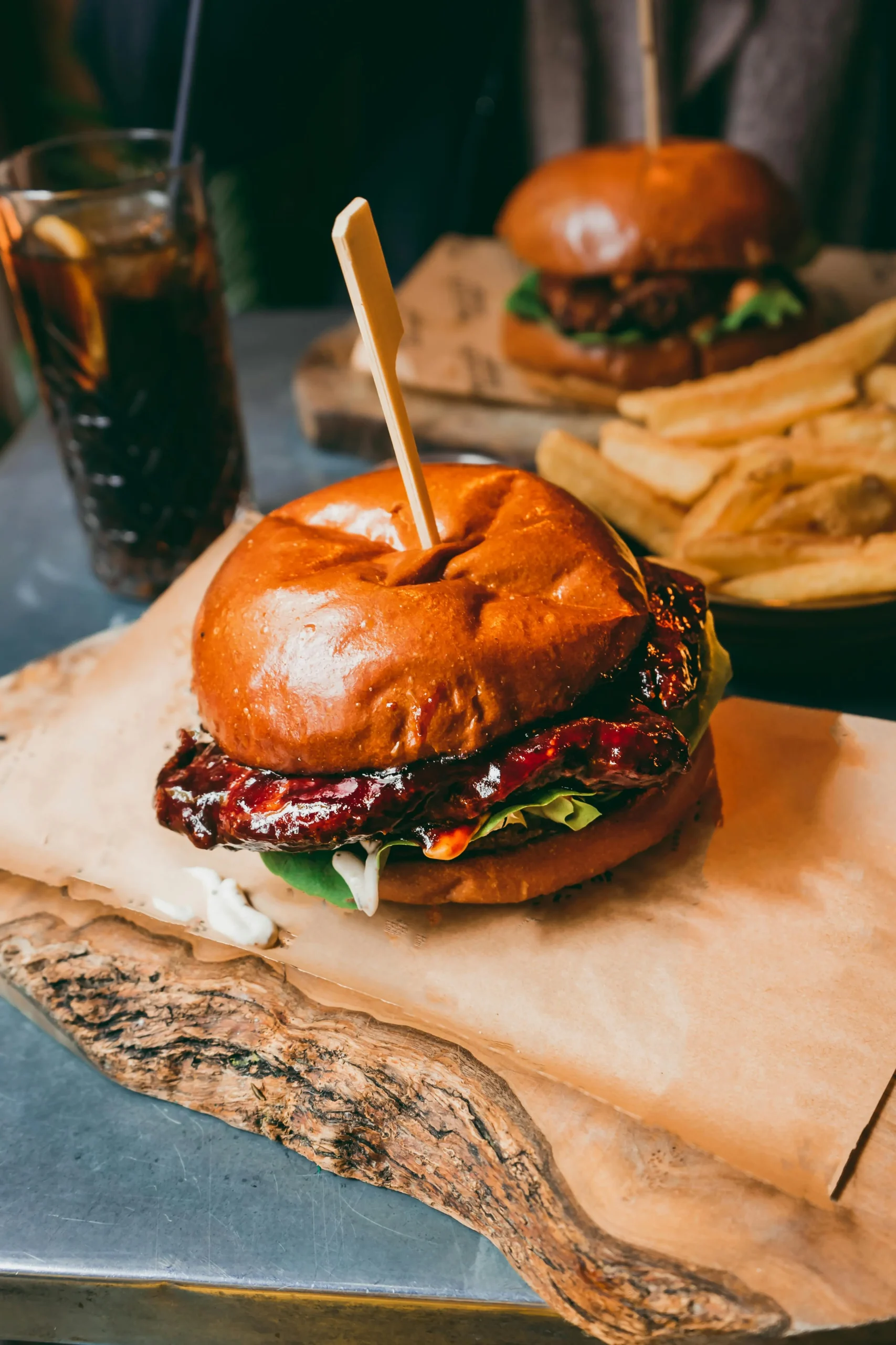 Primo piano di un hamburger al barbecue con lattuga e salsa in un panino lucido, tenuto insieme da uno spiedino di legno, servito su carta pergamena - ripreso con produzione foto e video professionale. Sullo sfondo, patatine e un bicchiere di soda sono leggermente sfocati.