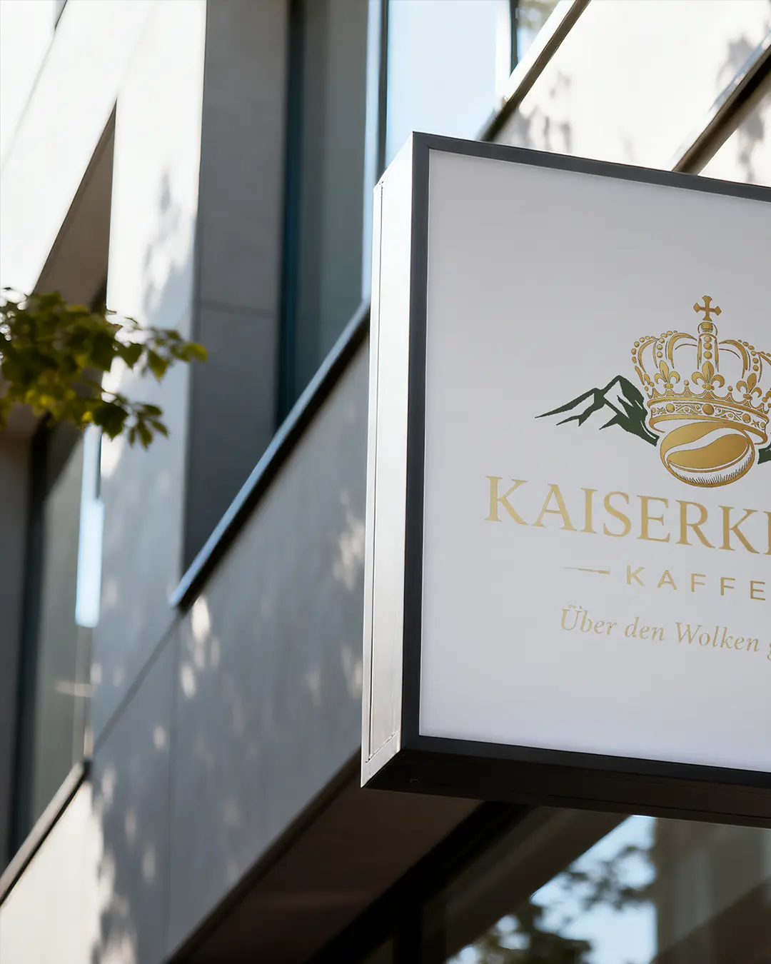 Un'insegna bianca con una corona dorata, un chicco di caffè e il logo della montagna KAISERK KAFFEE è appesa all'esterno di un edificio moderno. Sullo sfondo sono visibili un ramo d'albero e delle vetrate.