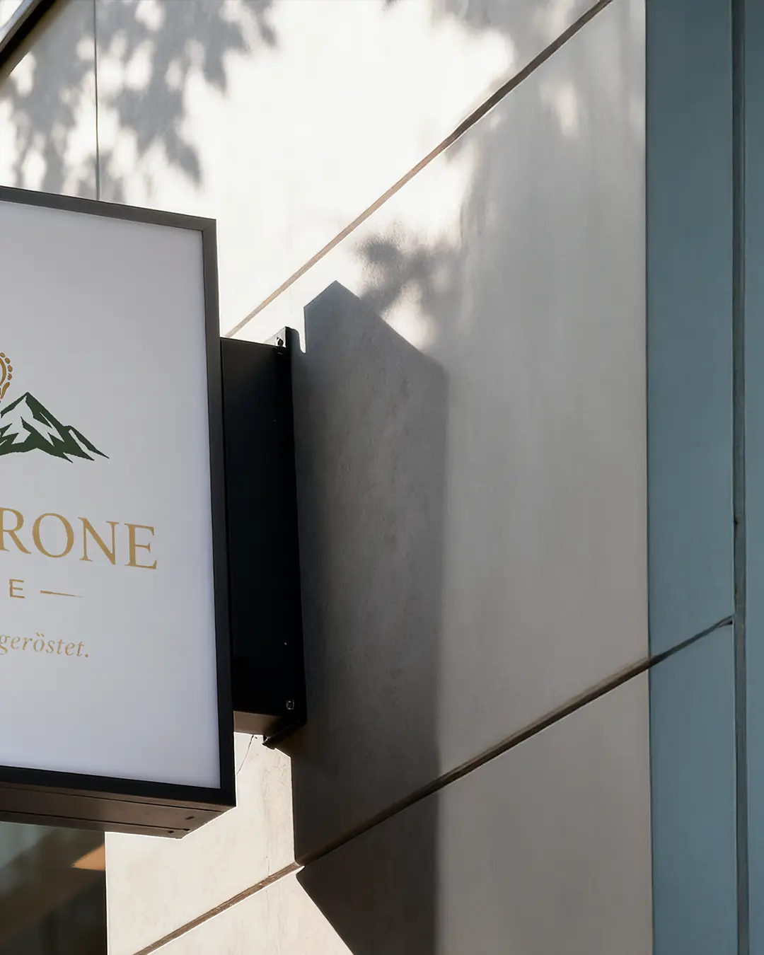 Un primo piano dell'esterno di un edificio presenta un'insegna parzialmente visibile con il logo di una montagna e la parola RONE in lettere dorate. La luce del sole proietta le ombre dei rami degli alberi sul muro grigio chiaro.