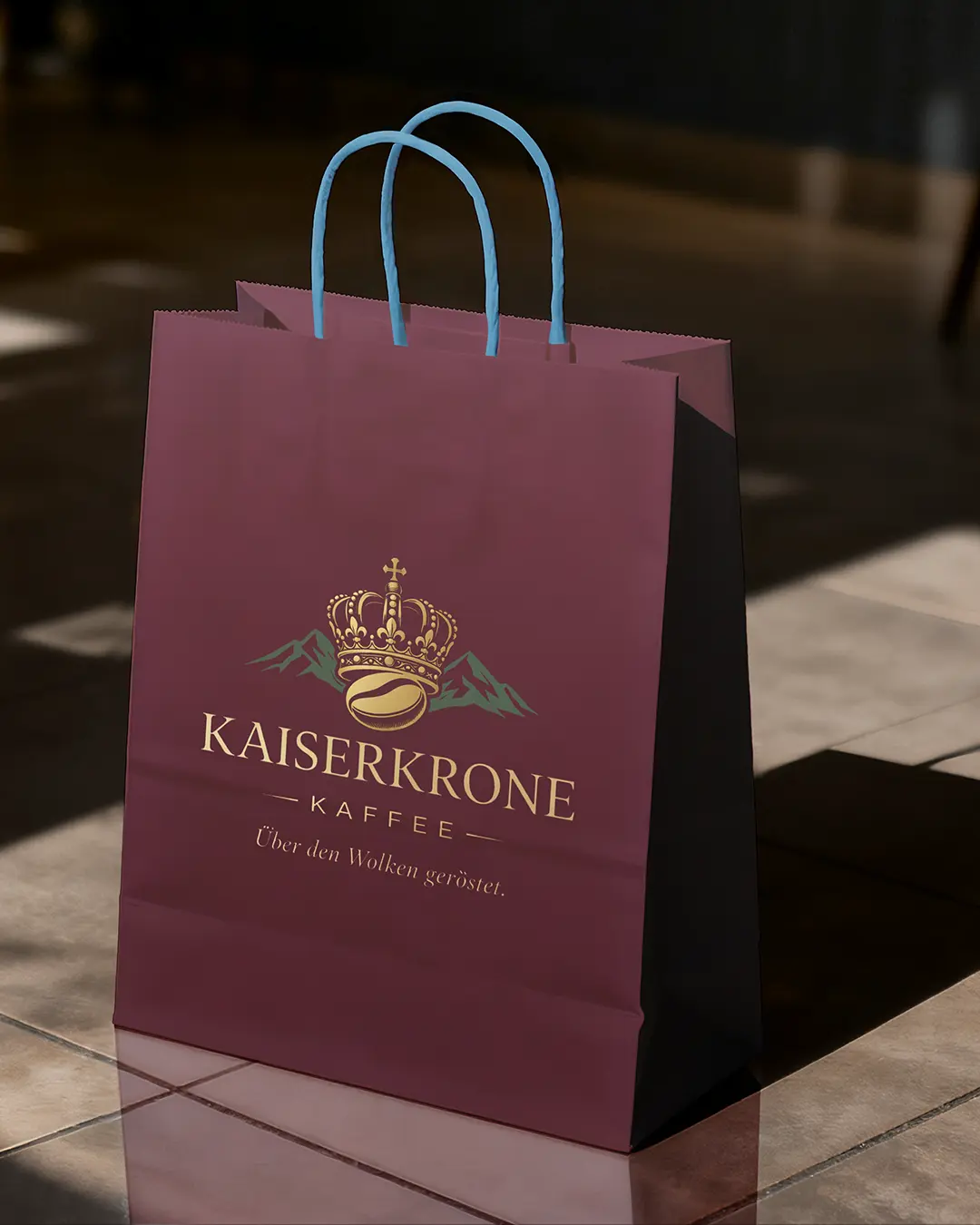 Una borsa della spesa marrone con manici azzurri è appoggiata su un pavimento piastrellato. La borsa presenta una corona dorata, montagne verdi e la scritta KAISERKRONE KAFFEE con una frase in tedesco.