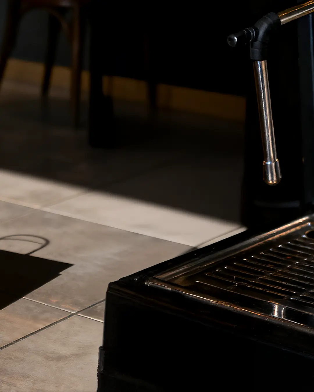 Primo piano di un lavandino di metallo lucido con un rubinetto alto in una stanza illuminata dal sole, che proietta ombre sul pavimento piastrellato. Sullo sfondo sono visibili parte di una sedia e di un tavolo.