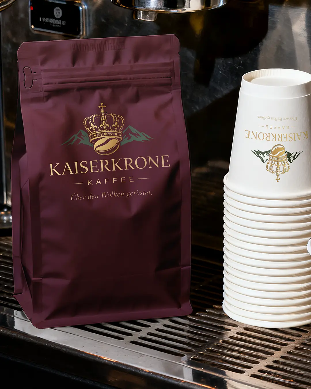 Un sacchetto marrone di caffè Kaiserkronen con logo oro e verde si trova accanto a una pila di bicchieri di carta bianchi a marchio Kaiserkronen sul bancone metallico di un bar.