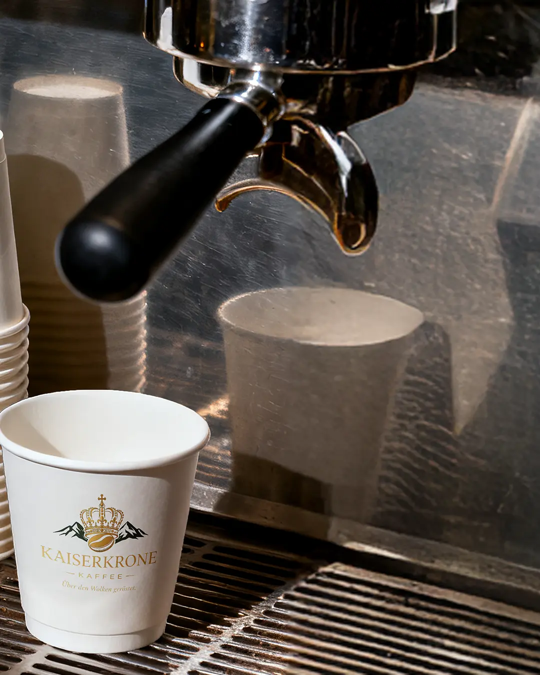 Primo piano di un portafiltro di una macchina da caffè sopra una tazza di caffè KAISERKRONE bianca, pronta per l'erogazione dell'espresso, con altre tazze di carta impilate nelle vicinanze e uno sfondo metallico.