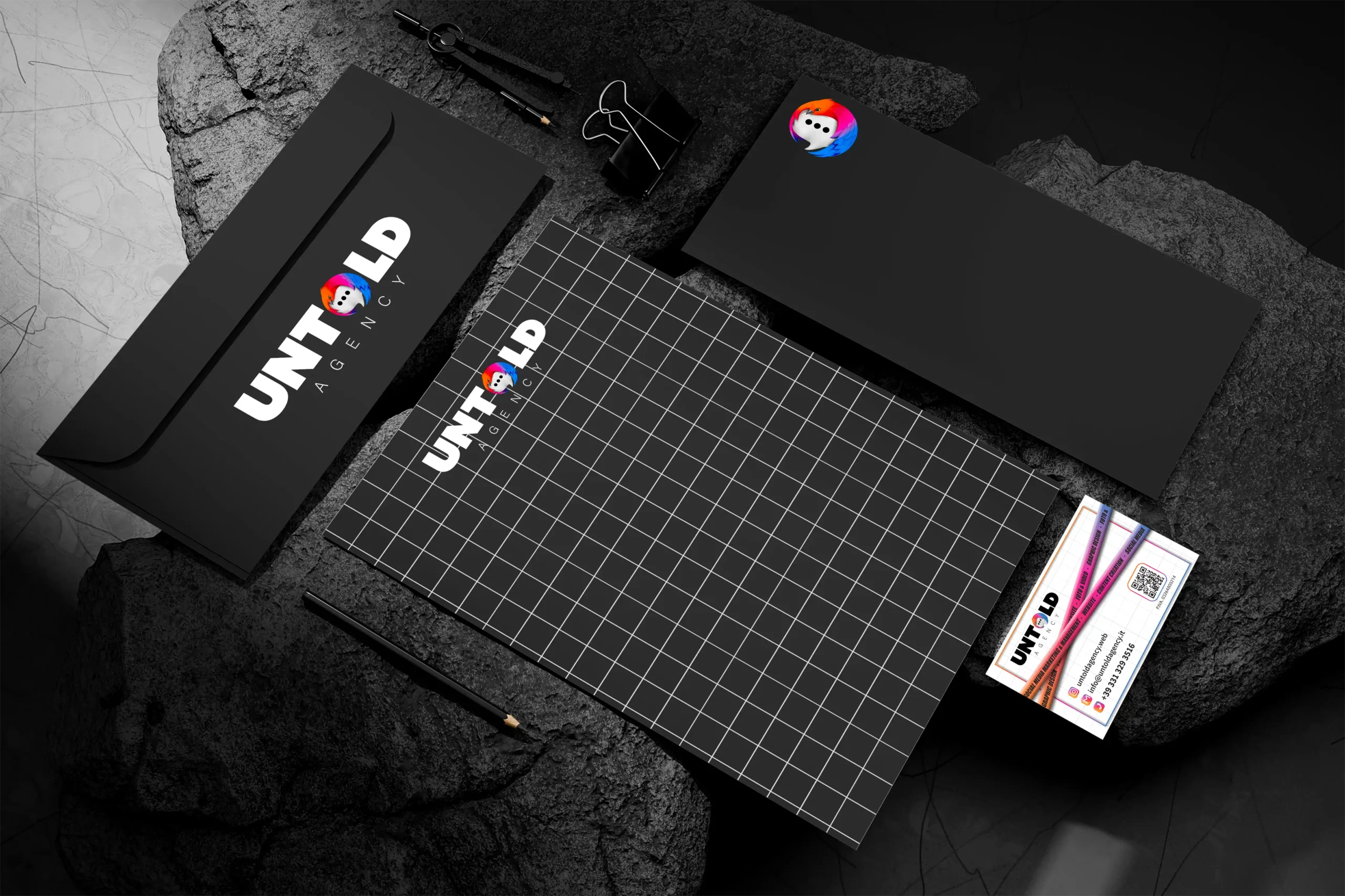 Un set di articoli di cancelleria di marca su una superficie di pietra scura, con buste nere, un blocco note a griglia, biglietti da visita e penne, tutti con il logo "UNTOLD AGENCY" e l'icona di una bolla di chat colorata.