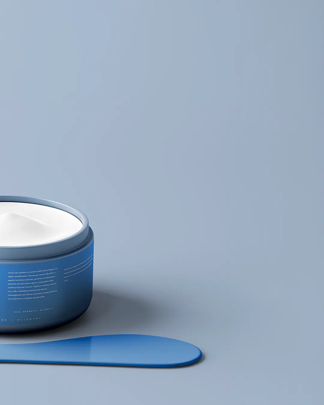 Un contenitore blu di crema bianca è aperto su una superficie liscia e azzurra. Davanti al contenitore si trova una spatola blu coordinata. Lo sfondo è minimalista e monocromatico.