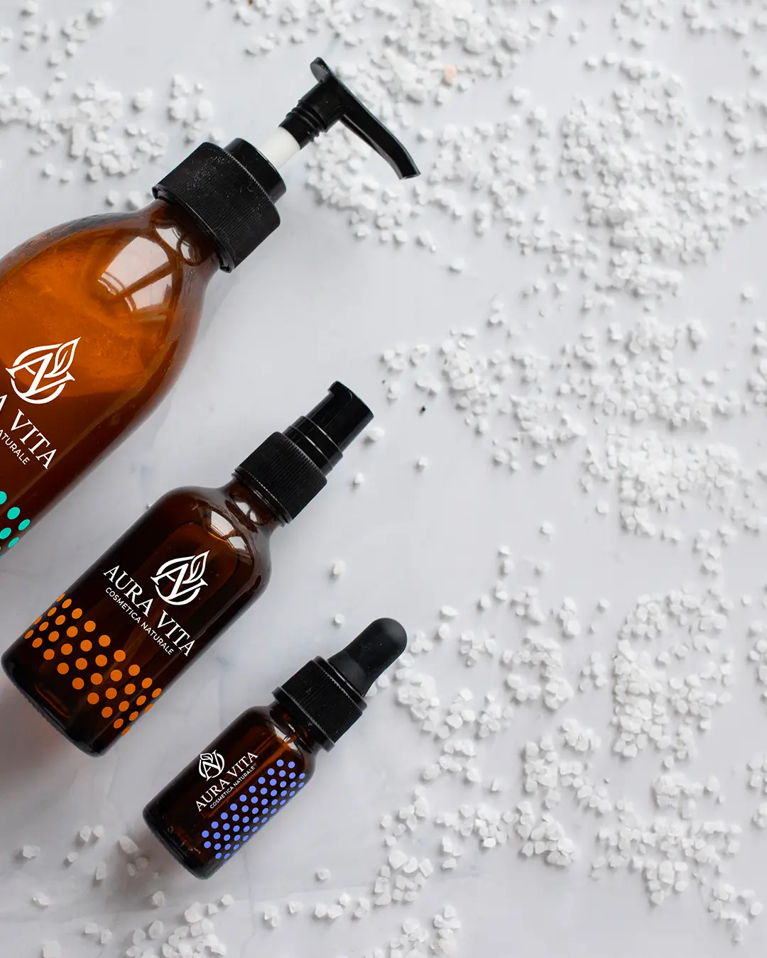 Tre flaconi marroni di prodotti per la cura della pelle Aura Vita con diversi erogatori (pompa, spray e contagocce) sono disposti su una superficie bianca disseminata di piccoli granuli bianchi.