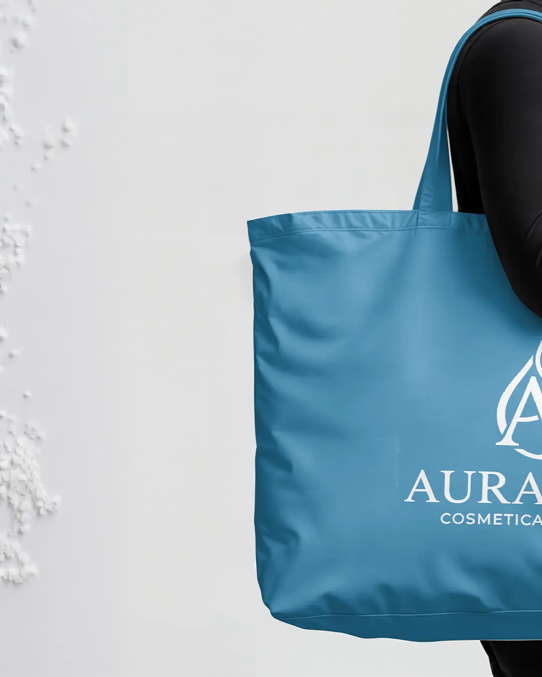 Una persona vestita di nero tiene in mano una grande borsa blu con la scritta "AURA COSMETICAL" e il logo "A" stilizzato, appoggiata a un muro bianco.