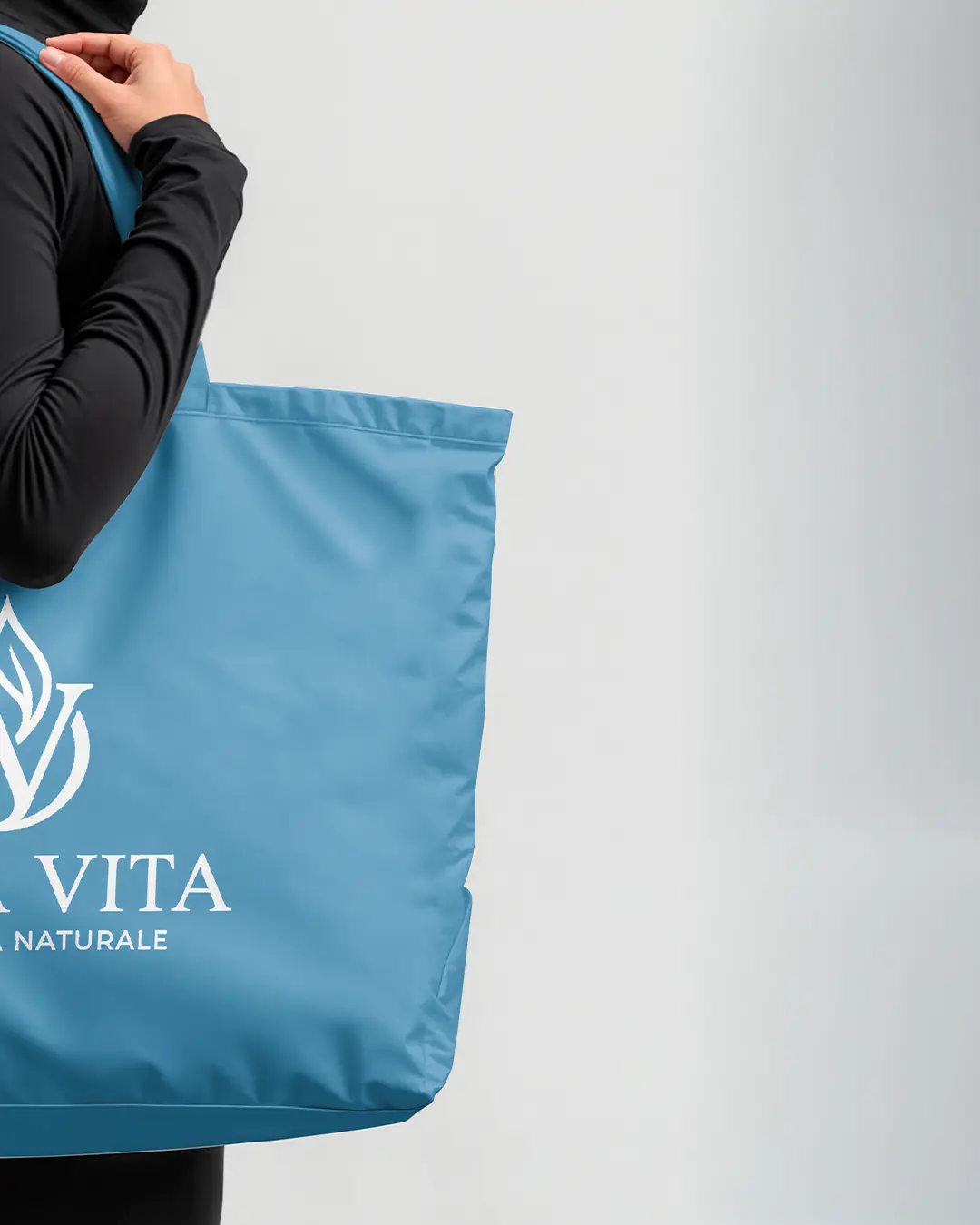 Una persona con un top nero a maniche lunghe tiene in mano una grande borsa blu con il logo di una foglia bianca e le scritte VITA e NATURALE parzialmente visibili. Lo sfondo è semplice e di colore chiaro.