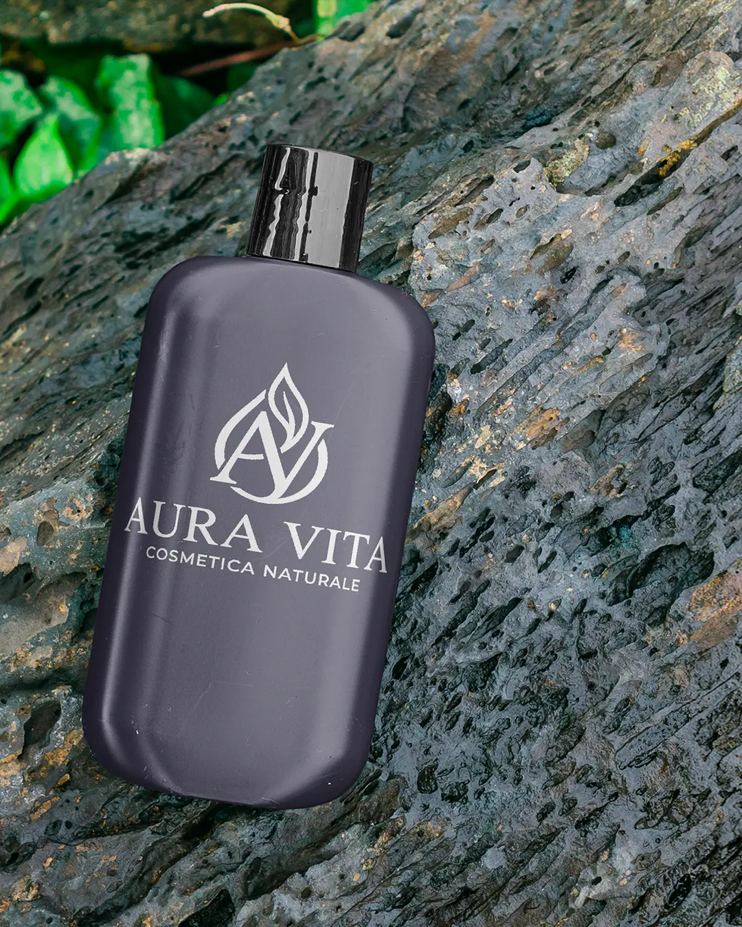 Una bottiglia scura con l'etichetta Aura Vita Cosmetica Naturale è appoggiata su una superficie rocciosa e strutturata, con un fogliame verde sullo sfondo.