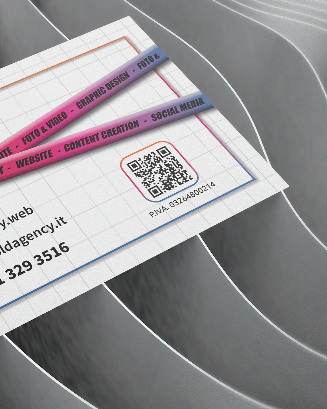 Primo piano di un biglietto da visita con strisce di testo colorate che elencano i servizi, un codice QR e informazioni di contatto, posizionato su una superficie con linee grigie curve e parallele.