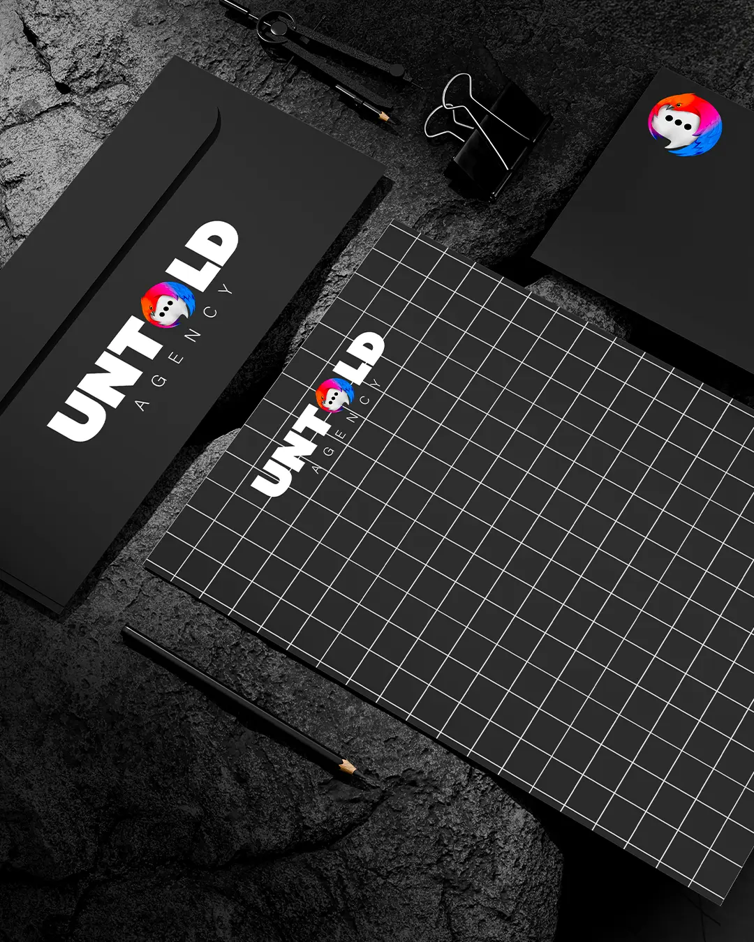 Un set di cancelleria nero con un quaderno a griglia, una busta, una matita, un fermaglio per raccoglitore e dei biglietti da visita, il tutto con il marchio UNTOLD AGENCY e un logo illustrato a colori con il volto di una persona. Lo sfondo è in pietra scura.