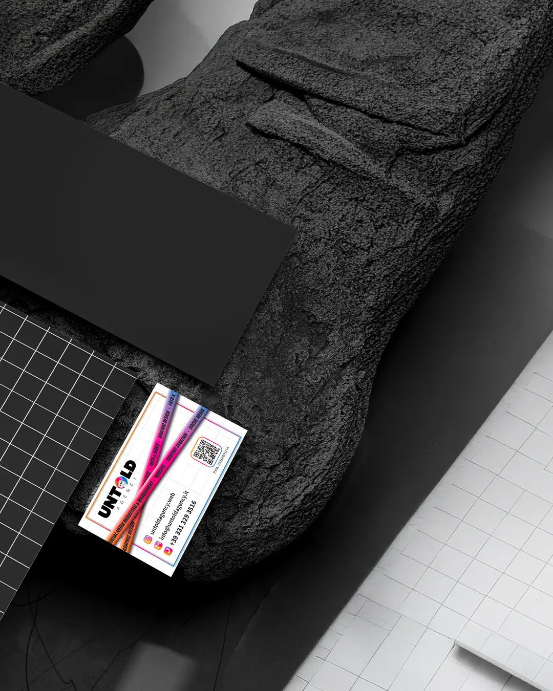 Un biglietto da visita colorato, con la scritta UNFOLD, è appoggiato su una superficie nera e strutturata, circondato da un taccuino nero, da una griglia e da un righello metallico.