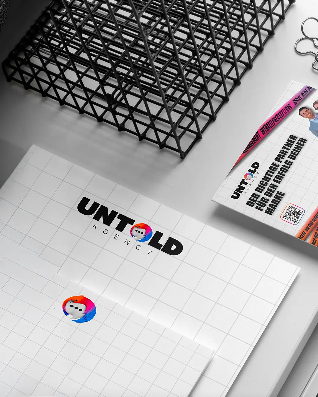 Una carta intestata e un biglietto da visita di Untold Agency sono appoggiati su una scrivania, accanto a un'agenda di metallo e a dei fermagli. Il logo presenta una faccia da cartone animato in un cerchio colorato e un testo nero in grassetto con la scritta UNTOLD AGENCY.