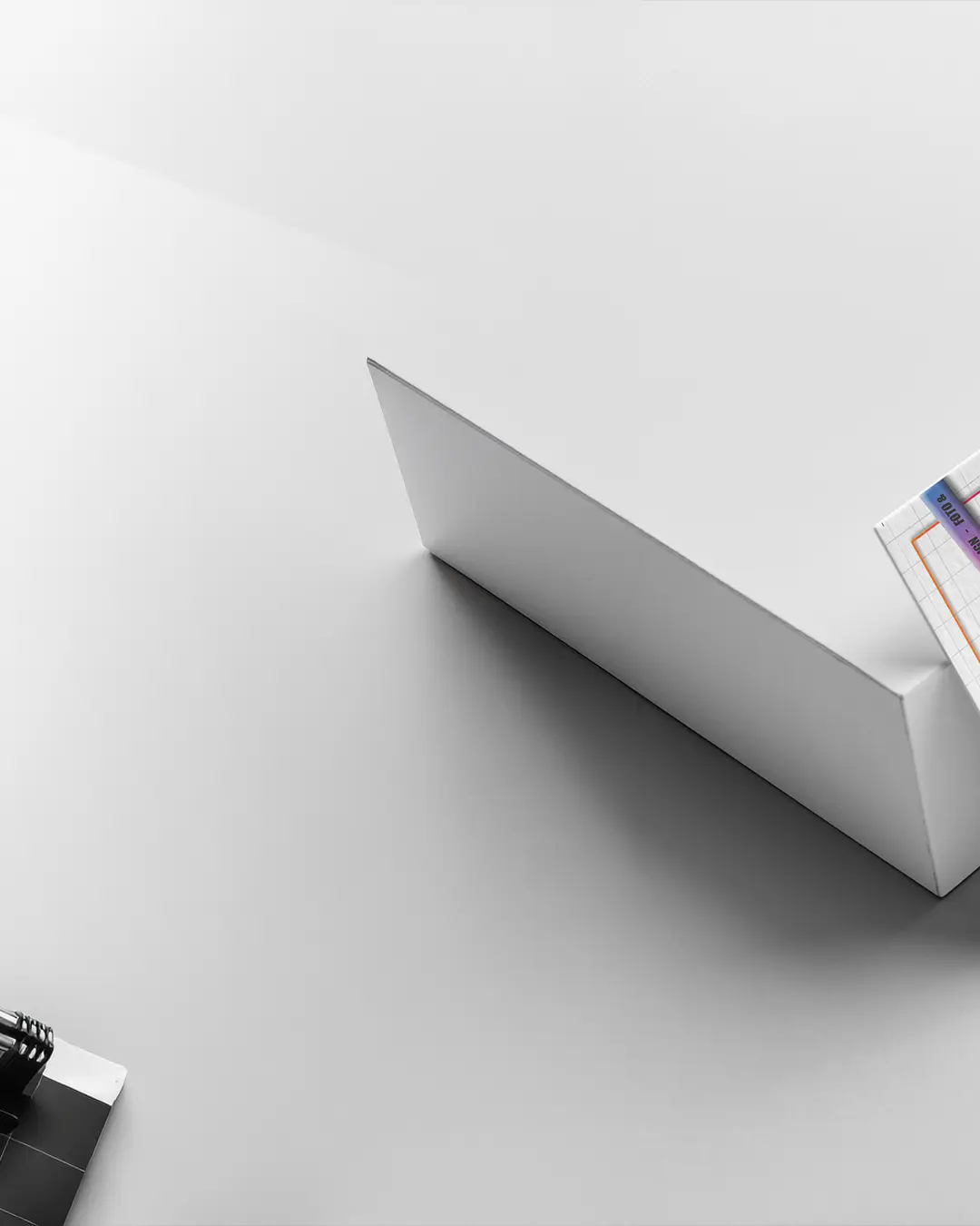 Spazio di lavoro minimalista con un computer portatile argentato chiuso su una superficie bianca. Sulla scrivania si trovano anche un angolo di una cartellina con carta millimetrata e un quaderno nero parzialmente visibile, che creano un'atmosfera pulita e organizzata.