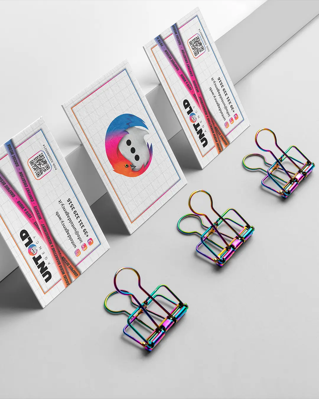 Quattro fermagli color arcobaleno sono disposti in fila accanto a quattro biglietti da visita, tre rivolti verso l'alto e uno verso il basso, il tutto su una superficie pulita di colore grigio chiaro con uno sfondo geometrico.