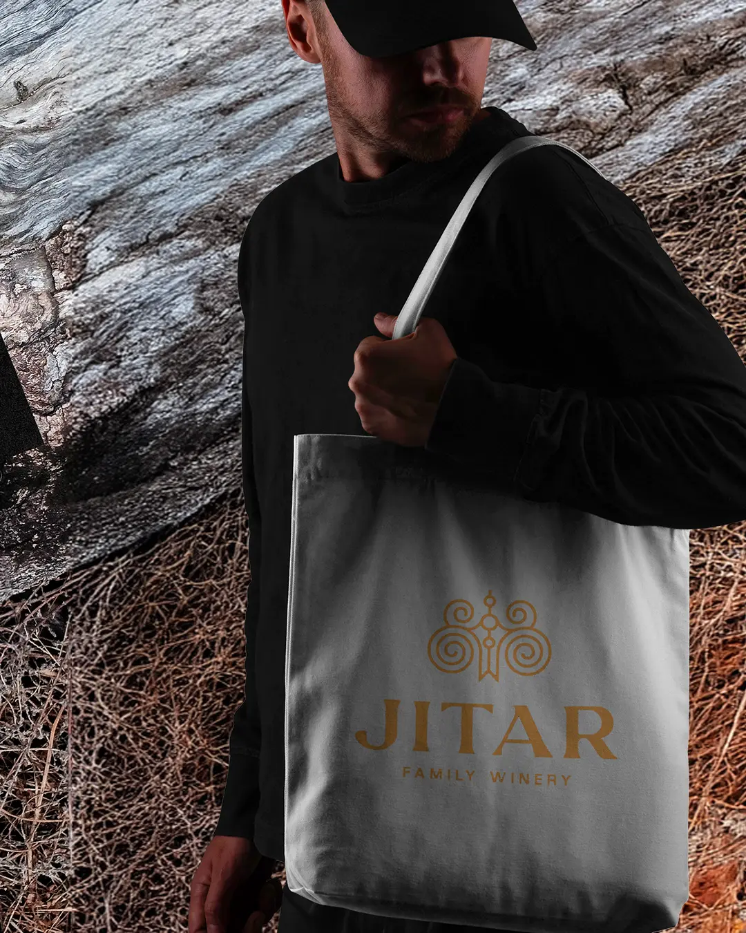 Una persona vestita di nero e con un berretto tiene in mano una tote bag bianca con il marchio JITAR FAMILY WINERY in oro. Lo sfondo è caratterizzato da legno strutturato ed erba secca.