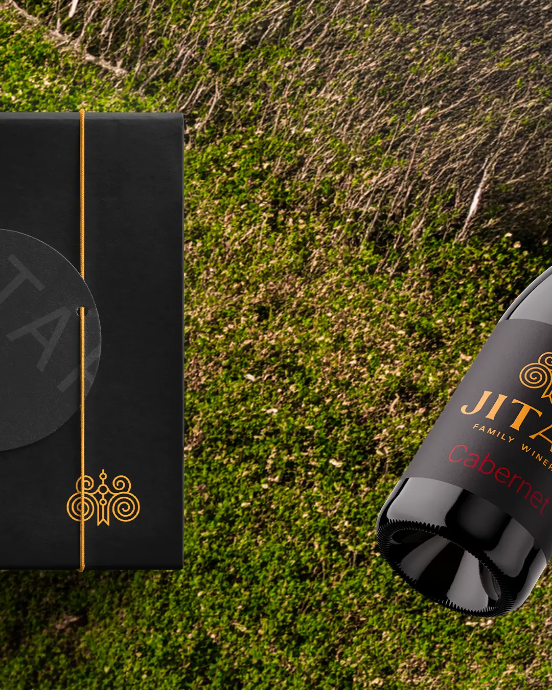 Una confezione regalo nera con accenti dorati e una bottiglia di vino cabernet Jitka giacciono su una superficie erbosa verde e strutturata.
