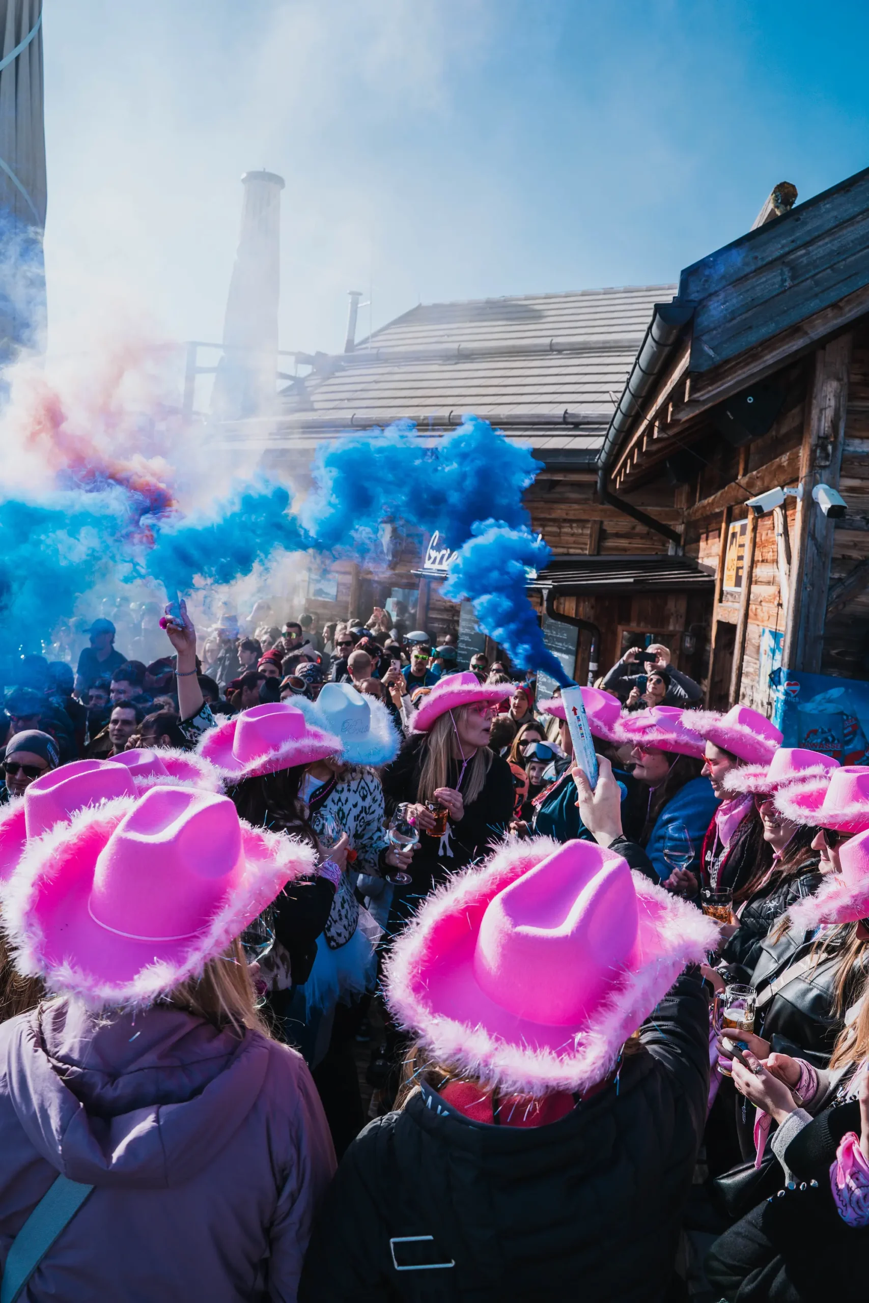 Una vivace folla all'aperto, molti dei quali indossano cappelli da cowboy rosa acceso, festeggia mentre nell'aria si diffondono razzi di fumo blu, circondati da edifici rustici in legno in una giornata di sole.