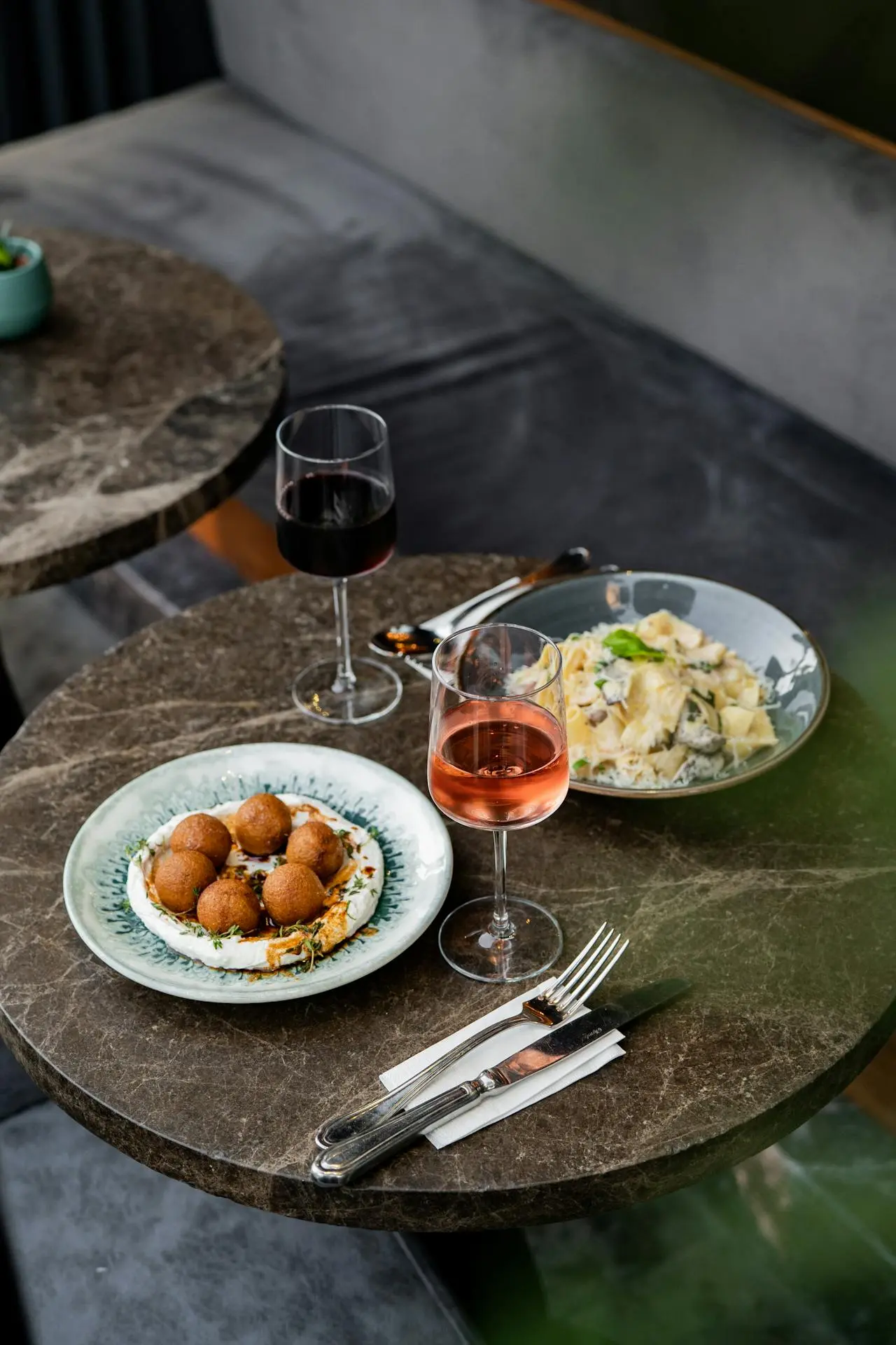 Un tavolo di marmo apparecchiato con due bicchieri di vino, uno rosso e uno rosato, accanto a un piatto di antipasti fritti e a un piatto di pasta cremosa. Le posate sono appoggiate su un tovagliolo accanto ai piatti.