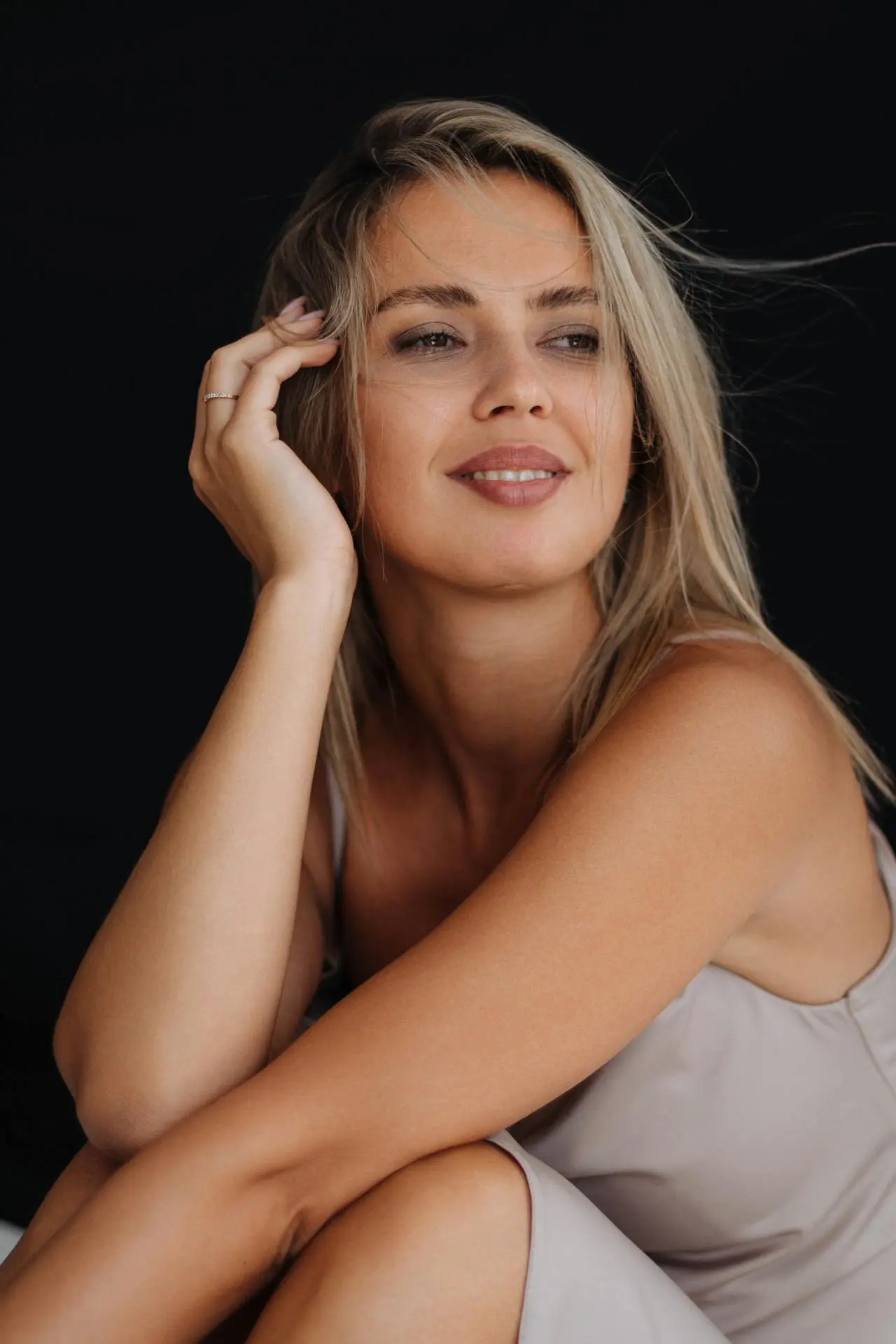 Una donna con i capelli biondi che indossa un top beige senza maniche è seduta con la mano appoggiata sul viso e sorride dolcemente su uno sfondo nero.