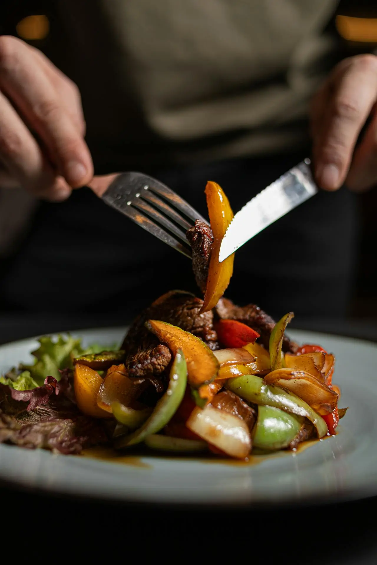 Una persona usa forchetta e coltello per tagliare un piatto con carne alla griglia e verdure colorate saltate, tra cui peperoni e cipolle, su un piatto con lattuga.