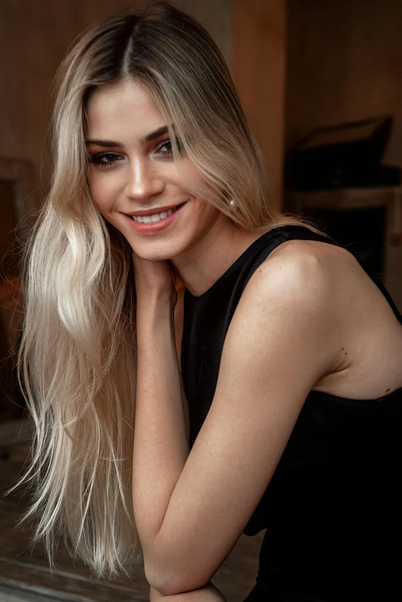 Una giovane donna dai lunghi capelli biondi e lisci e dalla pelle chiara sorride calorosamente all'obiettivo. Indossa un top nero senza maniche ed è seduta in un luogo chiuso con una luce naturale e soffusa.